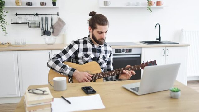 Smiling Caucasian man learning to play instrument using online lessons on laptop. Portrait of handsome bearded adult posing with six-string guitar while enjoying pleasant pastime at home.