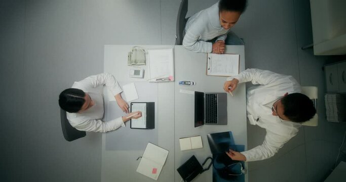 A doctor in a white coat examines an X-ray of a patient sitting in front of an open laptop at a table in a medical office, working in a clinic. The nurse is sitting across from him. View from above