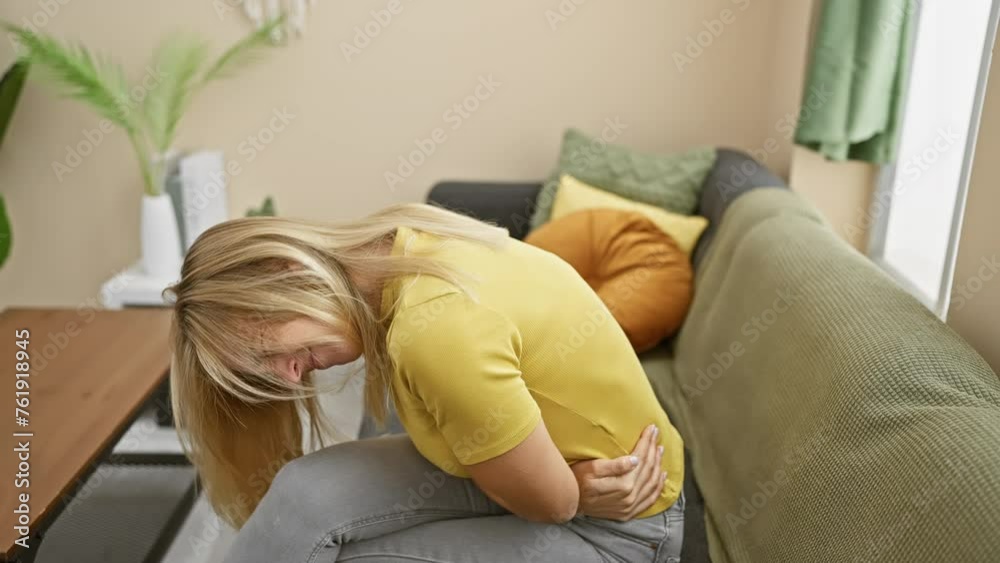 Poster Young blonde woman in pain at home, wearing t-shirt and clutching her stomach ill from a potentially serious health problem or abdominal issue