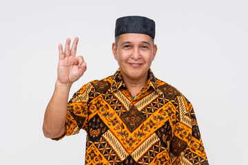 Smiling Indonesian man wearing a batik shirt and a kopiah hat making the OK gesture, isolated on...