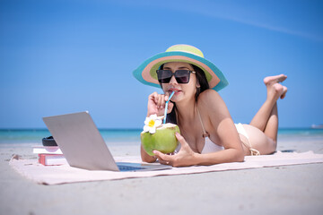 Happy sexy freelance woman wearing bikini sunglasses  and beach hat workout relaxed drinking coconut juice with work on laptop computer at beach in summer holidays.
