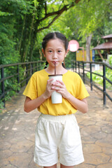 Portrait of Asian young girl kid holding paper cup and drinking cold water by straw with looking camera while standing in public garden. - 761951971