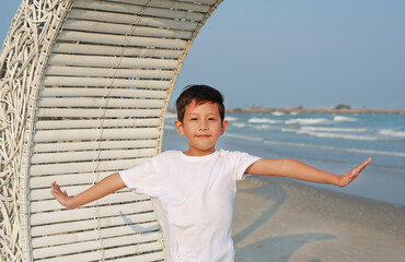 Portrait of smiling Asian boy open arms wide while standing on white bamboo wooden sofa bed with looking camera at the beach. - 761953327