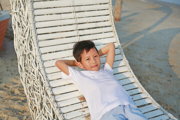 Portrait of happy Asian boy relax on white bamboo sofa bed and looking camera on the beach at summer holiday. - 761953344