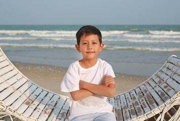 Portrait of smart Asian boy sitting with arms crossed on white bamboo wooden sofa bed with looking camera at the beach. - 761953374