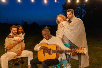 Obraz premium A group of friends enjoys a serene moment under the night sky, wrapped in blankets, as one plays the guitar, creating a cozy and musical outdoor experience. Cozy Nighttime Serenade at an Outdoor