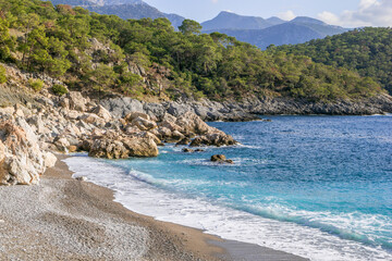 Beautiful small rocky beach with clear blue sea, waves and mountains on background. Oludeniz, Turkey.
