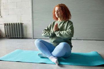 Fotobehang Pretty happy relaxed redhead female practicing yoga sitting in lotus posture on blue mat in gym, keeping hands in namaste gesture, doing stress relief techniques after hard work day at office © Anatoliy Karlyuk