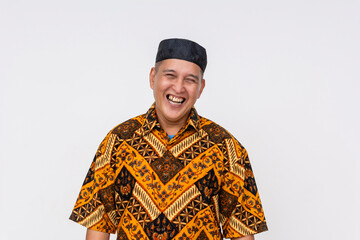 Portrait of a cheerful Indonesian man wearing a batik shirt and kopiah hat, laughing heartily isolated on a white background.