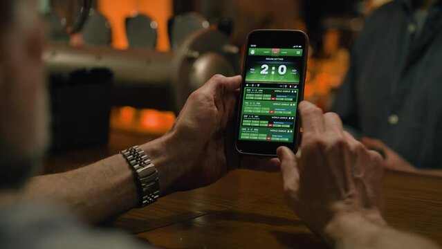 Close up of mature sports fan checking game score and bookmaker ratings in app using mobile phone. Caucasian man bets on football match online sitting at bar counter in pub at night. Gambling concept.