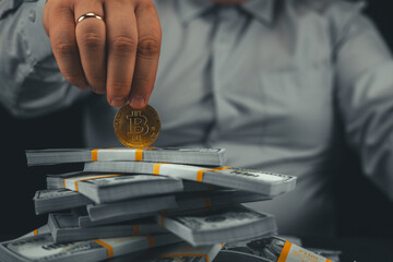 A man holds a bitcoin coin on a stack of stacks of dollars on a black background. A man in a...