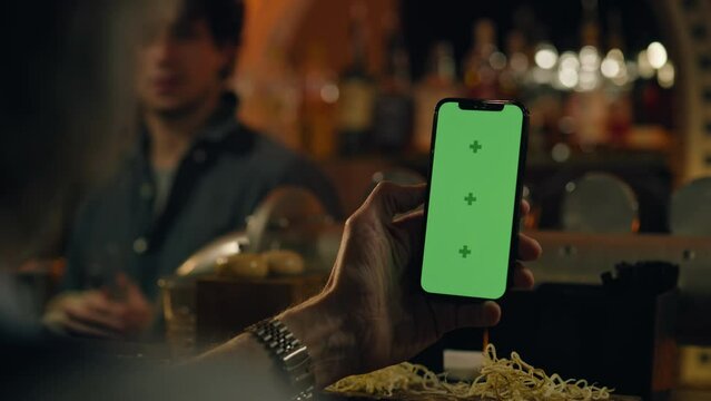 Close up of man holding phone with green screen, touching display. Sports fan watches bookmaker ratings online or surfs the Internet, talks with bartender sitting at bar counter in pub. Chroma key.