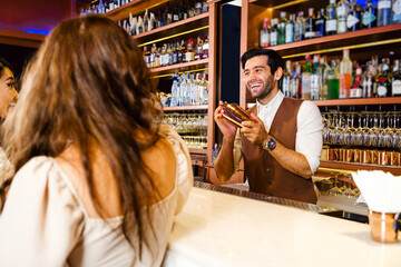 Caucasian professional bartender or mixologist making a cocktail for women at a bar. Attractive barman mixes liquor ingredients with a cocktail shaker with a smile. Bartender at night club restaurant. - 762032337