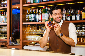 Caucasian professional bartender or mixologist making a cocktail for women at a bar. Attractive barman mixes liquor ingredients with a cocktail shaker with a smile. Bartender at night club restaurant. - 762032399