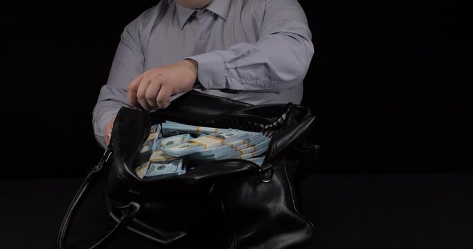 A man in a business shirt zips up a bag full of hundred dollar bills in front of him The man at the service was given a large bribe in a black bag A lot of money in a black bag on a black background.