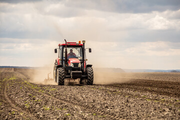 Powerful red tractor tills the soil on a vast farm with a dramatic cloudy sky overhead - obrazy, fototapety, plakaty