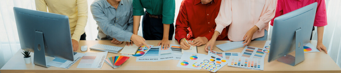 A top view cropped imaged of creative graphic team selects colors carefully from color wheel and palette on table at modern studio, surrounded by graphic documents and materials. Variegated.