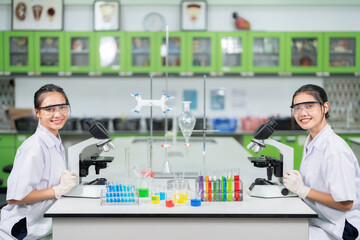 Naklejka na ściany i meble In a research lab, two Asian female scientists use a microscope to examine cells to produce a vaccine, smiling. On the table are experimental equipment, glass tubes, beakers, etc.