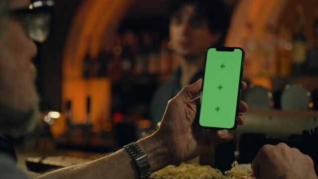 Mature man uses mobile phone with green screen, bets on football match online or surfs the Internet. Sports fan checks bookmaker ratings, talks with bartender sitting at bar counter in pub. Chroma key
