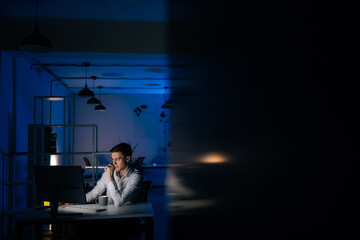 Remote view of serious young business man remote working overtime, learning online late at night in in dark room with neon light using desktop computer at workplace. Concept overwork and deadline.