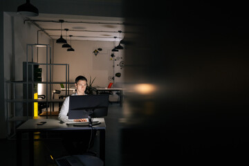 Remote view of focused young office worker male working at workplace on computer at late night in dark room. Successful entrepreneur checking finances on internet sitting at desk at evening.
