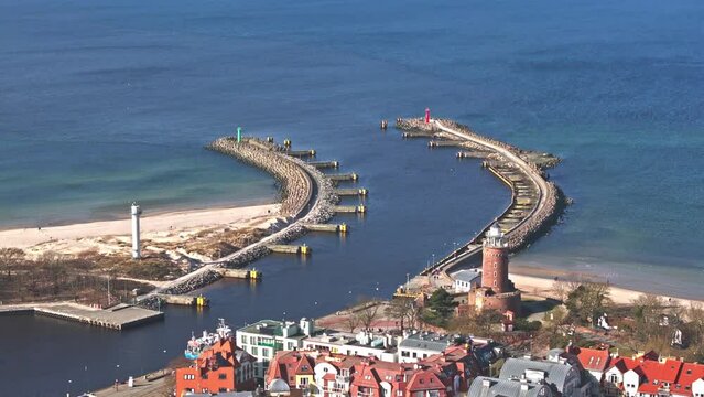 Captured by drone on a sunny February day in Kołobrzeg, the footage showcases port buildings, a lighthouse, beachgoers, and flying seagulls.