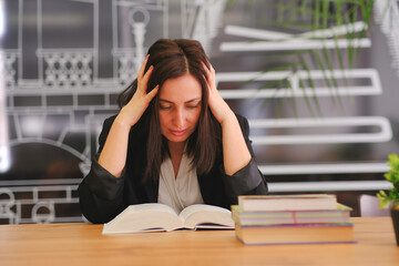 Young woman holding her head with her hands, serious. A woman holds her head with both hands, resting her elbows on a table in front of books, notebooks and documents