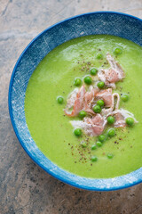 Fresh green pea soup served with bacon in a blue bowl, vertical shot on a beige granite surface, middle closeup