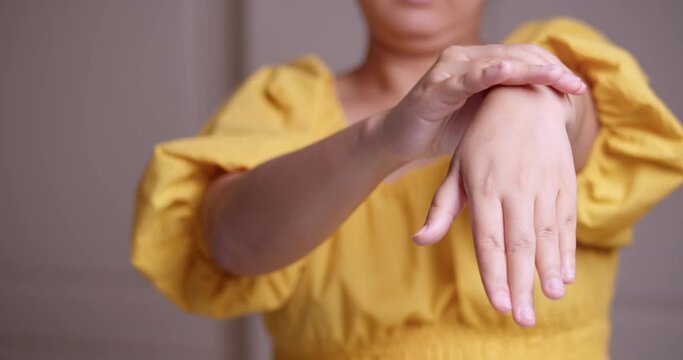 A close-up of a woman wearing a yellow dress is stretching her left arm while pinching and massaging herself from her wrist down to her fingers.