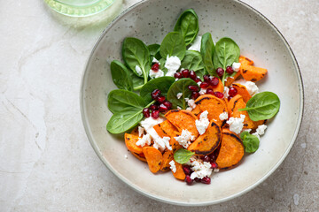 Plate with roasted sweet potato, fresh spinach, feta and pomegranate, horizontal shot on a beige stone background, high angle view