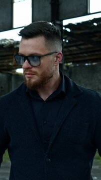Elegant man with a baseball bat outdoors. Portrait of a serious man in stylish glasses on the background of abandoned building. Vertical video