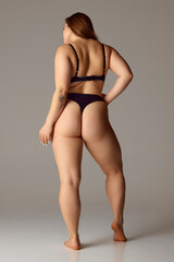 Full length rear-view portrait of young chubby woman posing in lingerie, black bikini against grey studio background. Concept of natural beauty, femininity, body positivity, dieting, fitness.