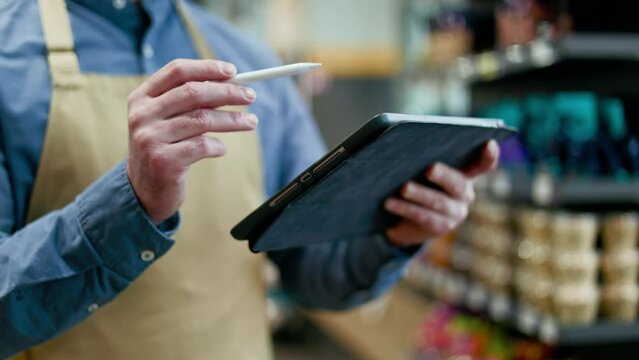Close up of unrecognizable retail manager in work clothes moving digital pencil across touch screen of modern tablet. Concerned person marking completed tasks from to do list on blurred background.