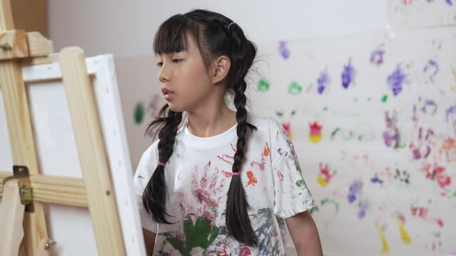 Playful student painted or draw canvas at stained wall in art lesson. Asian girl wearing white shirt with stained color while standing at stained wall with hand print. Creative activity. Erudition.