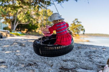 young girl toddler in red dress on a tyre swing on a sandy beach with soft toys