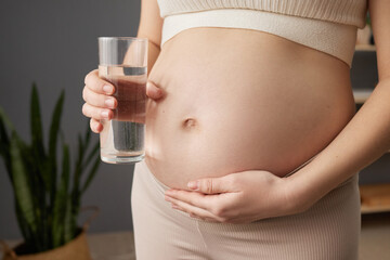 Unrecognizable pregnant woman with bare belly standing in home interior expectant mother touching her tummy holding glass of fresh water keeping water balance