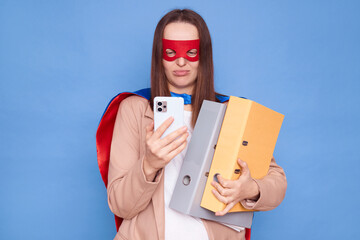 Unhappy woman wearing superhero costume standing isolated over blue background holding folders with...