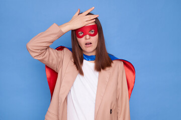 Fototapeta premium Displeased disappointed woman wearing superhero costume standing isolated over blue background having troubles showing facepalm gesture forgot something pleasant