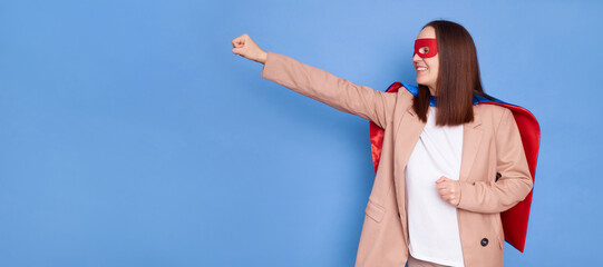 Side view of cheerful woman wearing superhero costume standing isolated over blue background looking at copy space for advertisement posing with raised fist © sementsova321