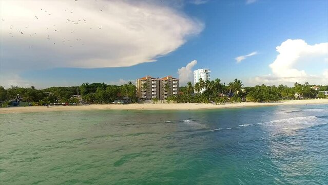 Approaching drone shot of the picturesque shoreline of Miami Beach, showing some luxurious hotels, beachgoers swimming and some birds flying in the sky, in Itea, Greece.