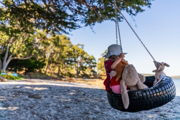 young girl toddler in red dress on a tyre swing on a sandy beach with soft toys