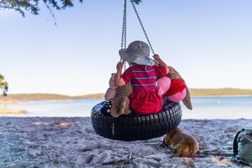 young girl toddler in red dress on a tyre swing on a sandy beach with soft toys - obrazy, fototapety, plakaty