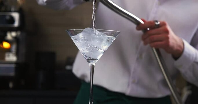 Professional bartender refrigerates Martini cocktail glass with ice cubes and cold tap water. Barman making cocktail at top bar. 