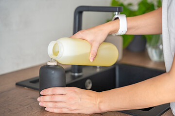 A woman pours soap or detergent from recycled packaging into a reusable bottle in kitchen. Eco-friendly lifestyle concept