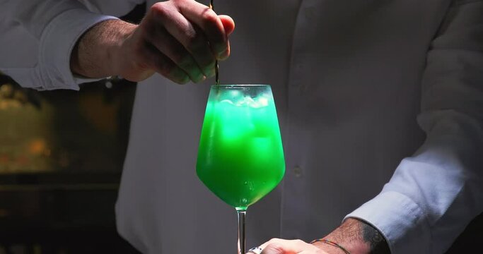 Close up of a professional bartender mixing an alcoholic green cocktail. Experienced barman mixes green alcoholic beverage. Bartender prepares cocktail. 4K slow motion.