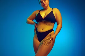 Foto op Plexiglas Cropped portrait of female plus-size model posing in dark bikini, underwear in yellow neon light against blue studio background. Concept of natural beauty, femininity, body positivity, diet, fitness. © Lustre