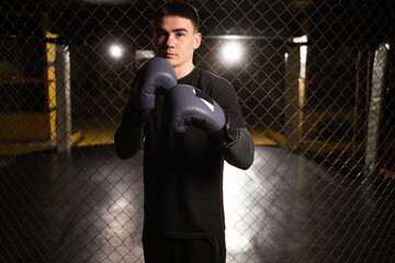 Young handsome boxer in boxing gloves standing near a metal cage in the gym looking at the camera