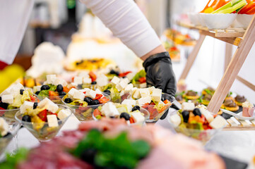 buffet where a person in gloves serves a variety of fresh salads. Colorful bowls contain ingredients like cheese cubes, olives, and chopped vegetables.