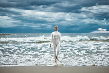 Hairless girl with alopecia in white futuristic suit comes out of cold restless sea on sandy beach,...