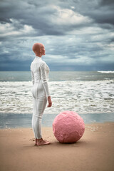 Hairless performer girl with alopecia in white futuristic suit stands on sea beach with pink sphere...